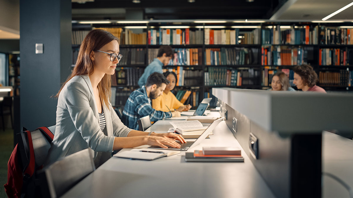 University Library: student Sitting at the Desk, Uses Laptop, Writes Notes for the Paper, Essay, Study for Class Assignment. Diverse Group of Students Learning, Studying for